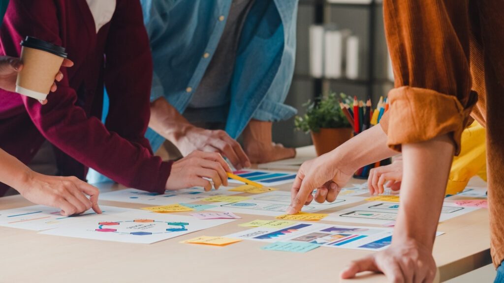 Grupo de pessoas usando roupas coloridas estão debruçadas em cima de uma mesa, analisando gráficos, estratégias e processos ágeis.