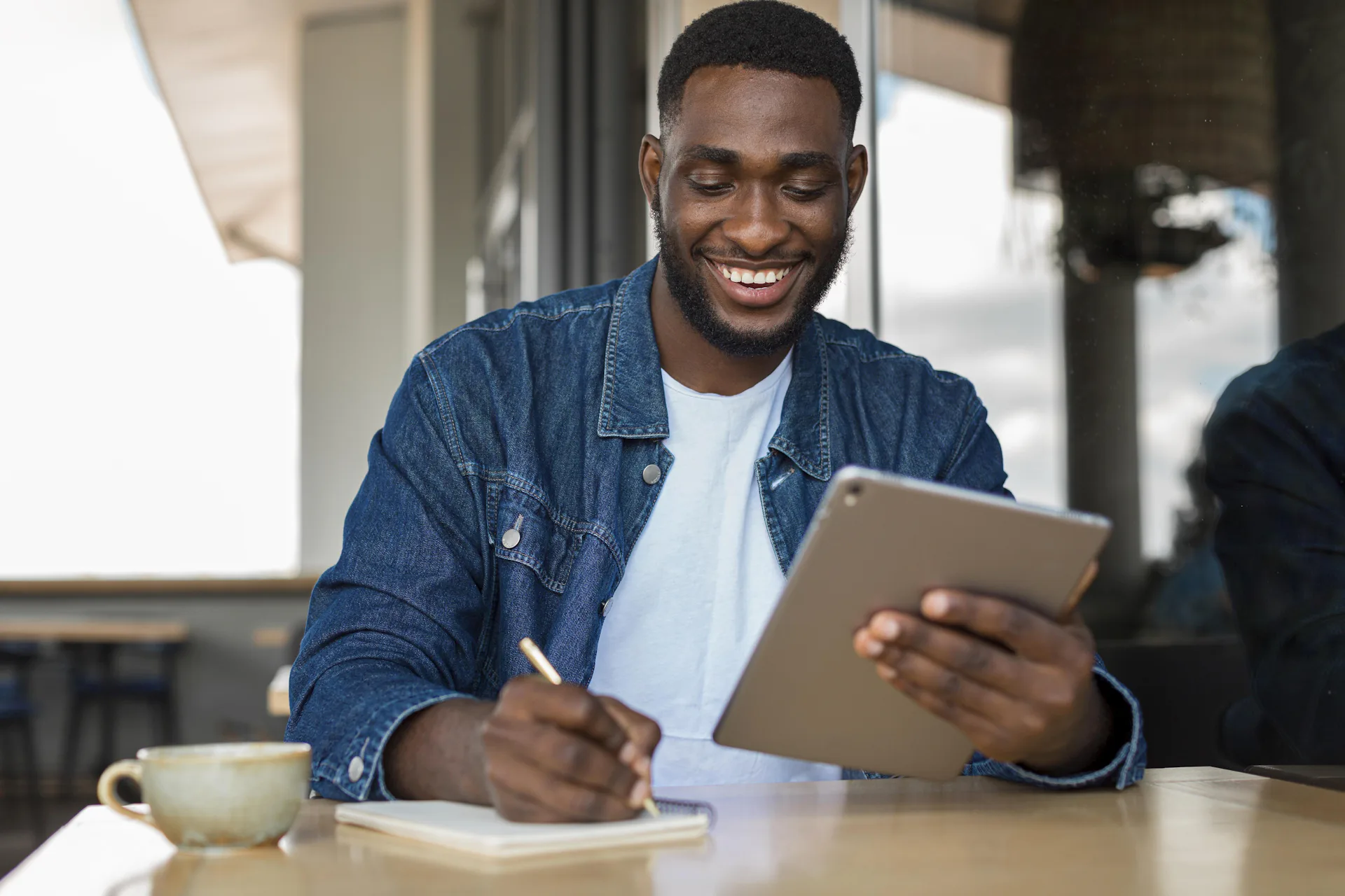 homem negro com cabelo curto e barba, vestindo uma camiseta branca por baixo de uma camisa jeans, está sentado em uma mesa escrevendo em um papel, segurando um tabblet e com uma xícara de café no canto esquerdo