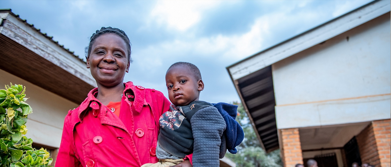 Uma mulher sorri enquanto segura seu filho nos braços, diante de uma construção simples sob um céu nublado. A expressão serena transmite esperança e força. A cena representa o impacto humano dos programas da Fundação Elizabeth Glaser de AIDS Pediátrica — vidas transformadas por cuidado, acolhimento e tecnologia a serviço da saúde.