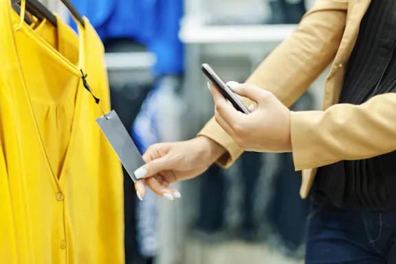 Mulher segurando smartphone e escolhendo roupas em uma loja física.