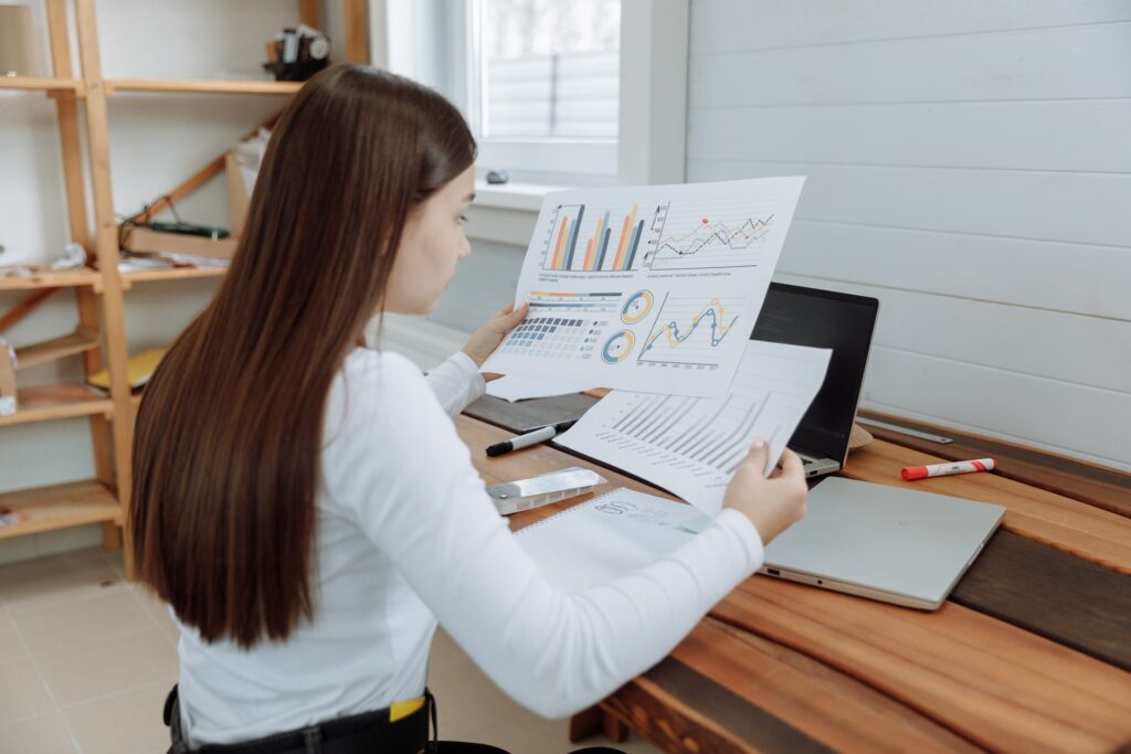 Mulher com blusa branca de manga comprida e calça jeans preta está sentada à mesa de trabalho analisando vários papéis com gráficos e indicadores.