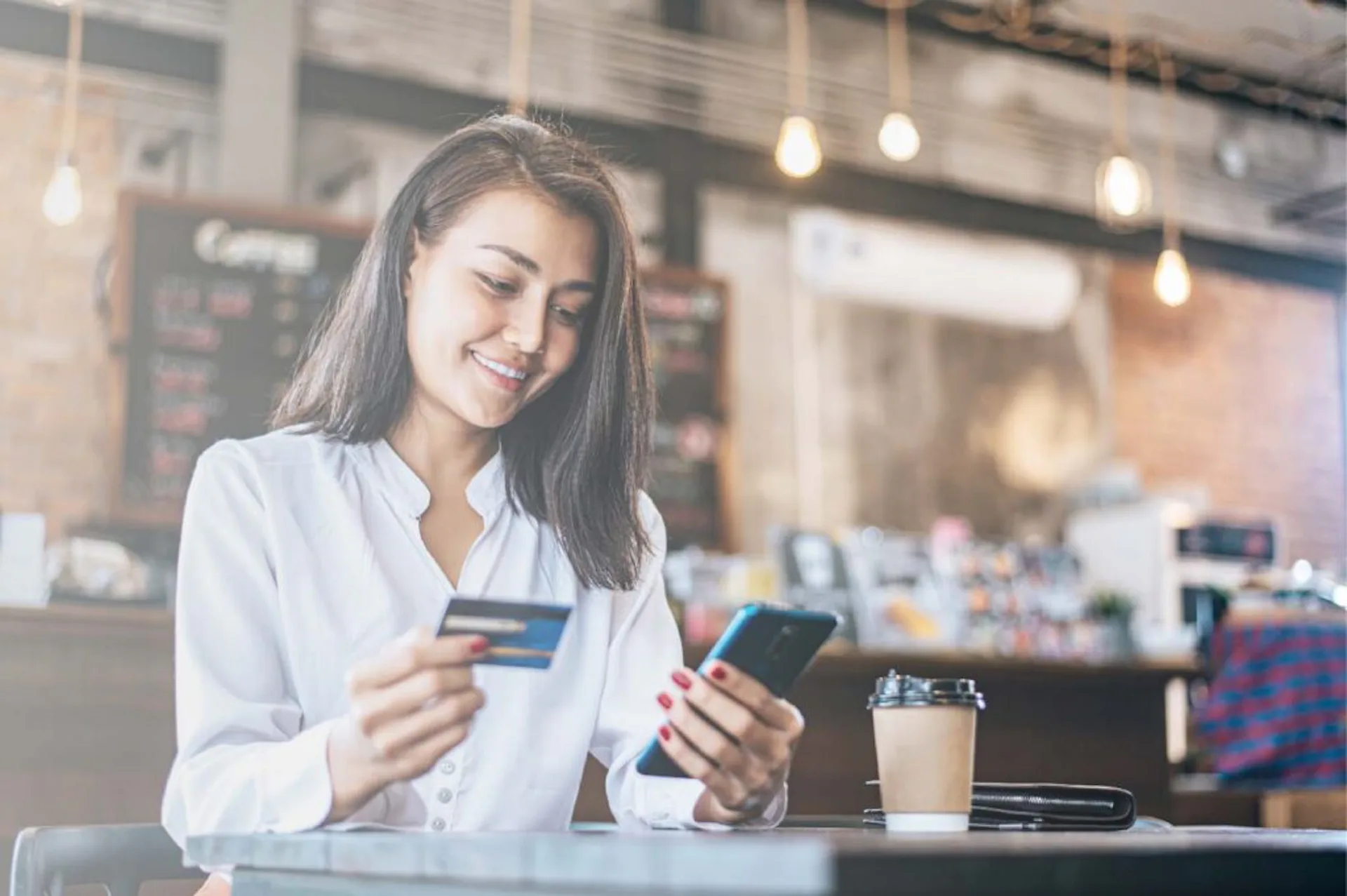 Mulher mexendo no celular e olhando o cartão de crédito em uma cafeteria.