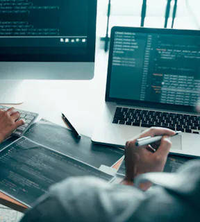 A imagem destaca duas telas de computador com códigos escritos na tela. Vê-se duas mãos, de diferentes pessoas, apontando para a tela e segurando objetos, como lápis, papel e caneta