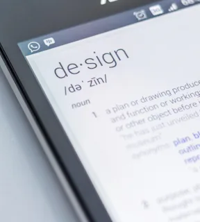 Detalhe da ponta de um celular, com a tela acessa e o significado da palavra Design em inglês como resultado de busca do aparelho.