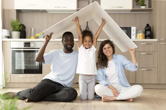 Foto de uma família com pai, mãe e criança ao meio, sentados na cozinha de sua casa, segurando um objeto simulando um telhado 