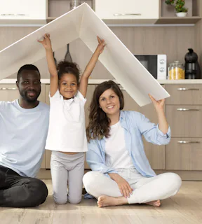 Foto de uma família com pai, mãe e criança ao meio, sentados na cozinha de sua casa, segurando um objeto simulando um telhado 