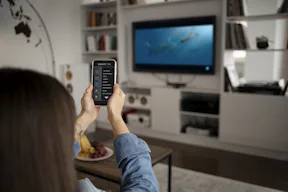 Photo of a woman holding a cellphone with a living room scene in the background focusing on the TV