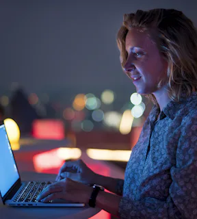 Mulher em pé mexendo em seu notebook sobre a mesa, em um ambiente aberto  de noite com as luzes da cidade ao fundo