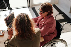 Uma mulher loira sentada à mesa de trabalho com uma mulher de óculos e blazer rosa em cadeira de rodas. À frente delas tem um notebook aberto em cima da mesa