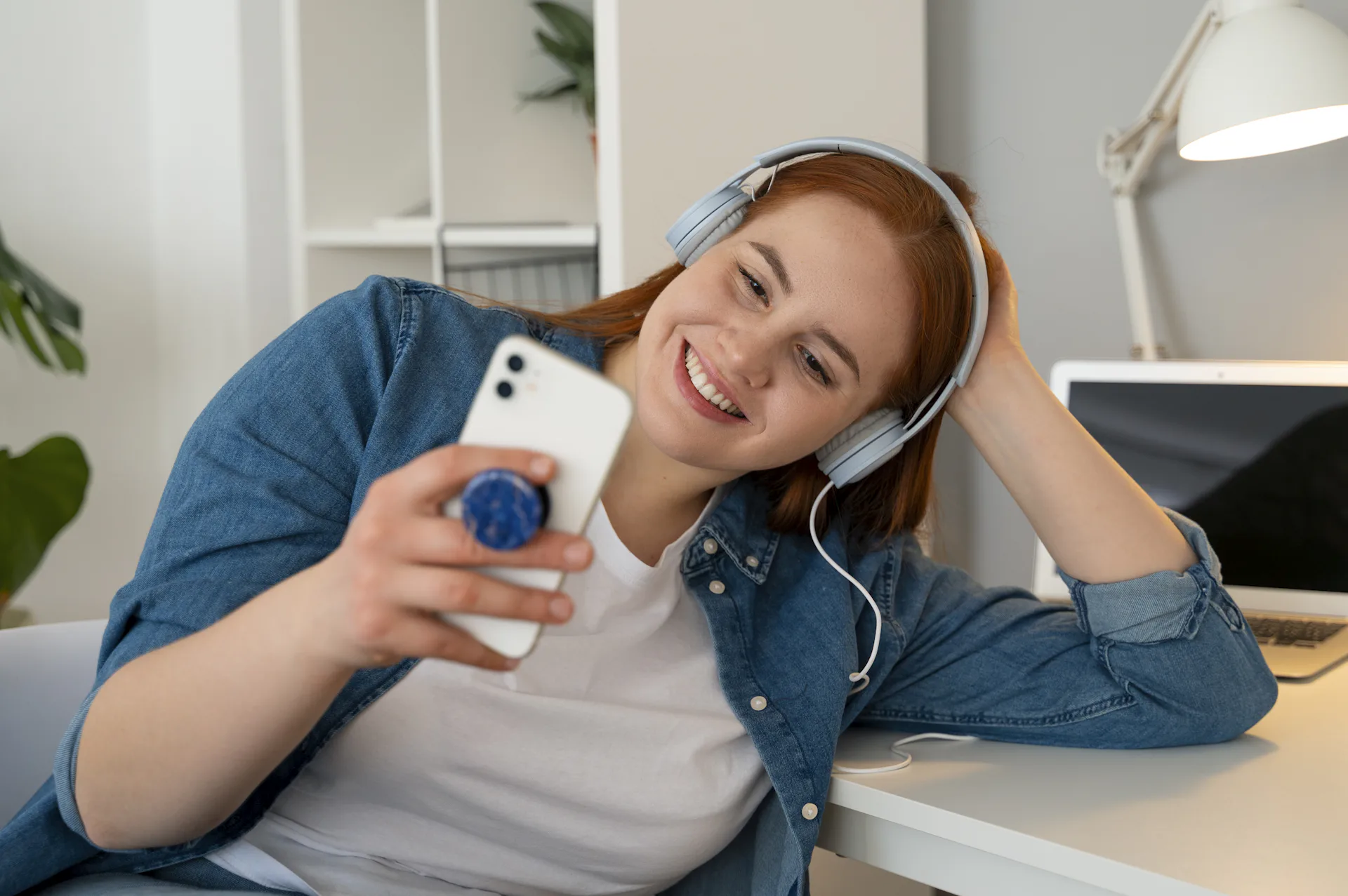 Foto com uma menina ruiva com fones de ouvido branco sentada segurando o celular com a mão esquerda e com a mão direita apoiando a cabeça 
